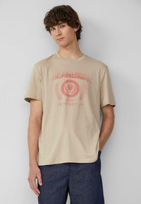 T-shirt en coton beige à manches courtes, avec un imprimé graphique rouge et du texte. Encolure ronde et coupe décontractée.