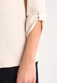 Blusa color crema con finitura testurizzata, dotata di maniche arrotolate fissate da bottoni. Tessuto liscio che si estende fino all'orlo sopra dei jeans scuri.