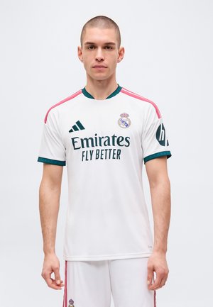 Giovane uomo con i capelli corti che indossa una maglia bianca del Real Madrid con strisce rosa e loghi degli sponsor, in piedi davanti a uno sfondo neutro.