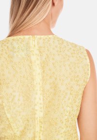 Gelbes ärmelloses Kleid mit zarter Blumenstickerei, mit einem sichtbaren Reißverschluss auf der Rückseite und einem strukturierten Stoff.