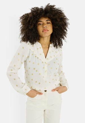 Femme aux cheveux bouclés portant un chemisier blanc à motifs d'étoiles dorées et un pantalon blanc, mains dans les poches, fond neutre.