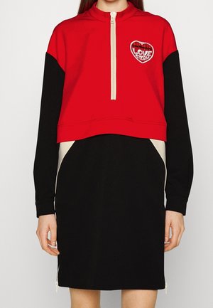 Robe sweat-shirt courte rouge et noire avec un col zippé, un patch en forme de cœur et des panneaux latéraux contrastants. Fabriquée en tissu doux avec une texture lisse.