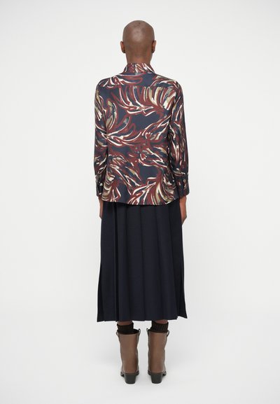 Langærmet bluse med et flerfarvet bladmønster på mørkt stof, parret med en mørk plisseret nederdel og knæhøje brune støvler.