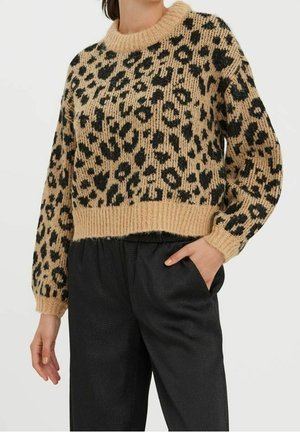 Femme portant un pull tricoté beige et noir à imprimé léopard avec poignets et ourlet côtelés, associé à un pantalon noir taille haute, une main dans la poche.