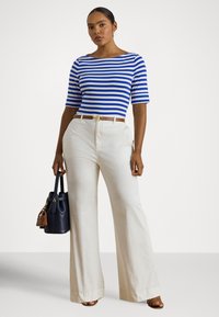 Woman wearing blue and white striped top, high-waist white wide-leg pants, holding navy handbag, standing against plain background.
