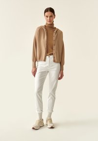 Cardigan beige con bottoni sopra un dolcevita marrone, abbinato a pantaloni bianchi arrotolati e sneakers beige con dettagli color nocciola.