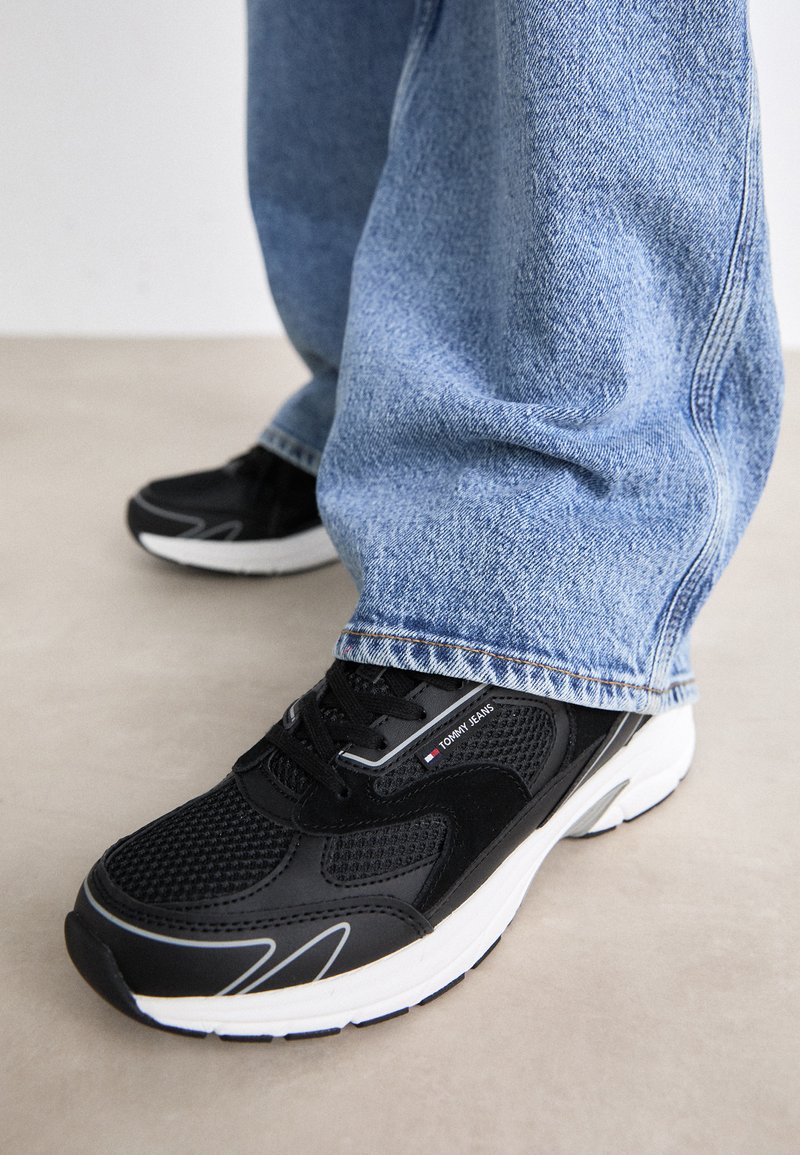 Baskets en mesh noir avec des semelles blanches, présentant un accent de logo et des lignes de conception courbées, associées à un jean en denim bleu clair.