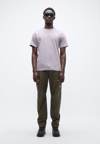 Ανοιχτό μοβ μπλουζάκι με λογότυπο, χακί παντελόνια και μαύρα μποτάκια. Το μοντέλο φοράει σκούρα γυαλιά ηλίου και στέκεται μπροστά σε απλό φόντο.