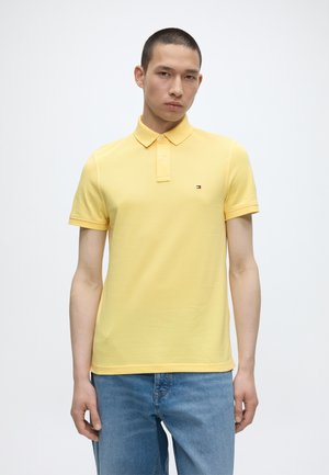 Hombre con el pelo corto que lleva una camiseta polo amarilla y vaqueros azules, de pie frente a un fondo blanco liso.