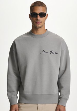 Homme portant un sweat-shirt gris col rond avec le texte "Mira Paris" sur la poitrine et des lunettes de soleil rectangulaires noires sur fond blanc.