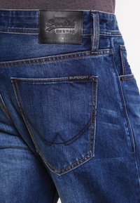 Tmavě modré džíny s úzkým střihem, které mají koženou nášivku na pase s označením "Superdry" a kontrastní prošívání.