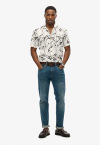 Camisa blanca de botones de manga corta con estampado de bambú negro; combinada con jeans azules remangados en el puño; cinturón y zapatos marrones.