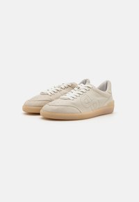 Beige lærersneakers med rund tå, hvite snøringer, perforerte detaljer og en lys brun gummisåle. Glatt tekstur og minimalistisk design.