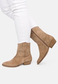 Botines de ante tostado con patrones bordados en espiral, tacón bajo de madera y detalle de cremallera lateral. Llevados con pantalones ajustados blancos.