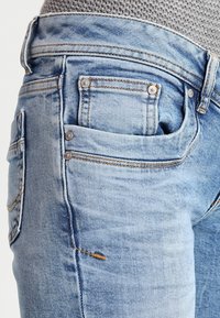 Jasnoniebieskie dżinsy z efektownym wyblakłym wykończeniem. Posiadają wiele kieszeni oraz metalowe guziki, prezentując klasyczny pięciokieszeniowy krój.