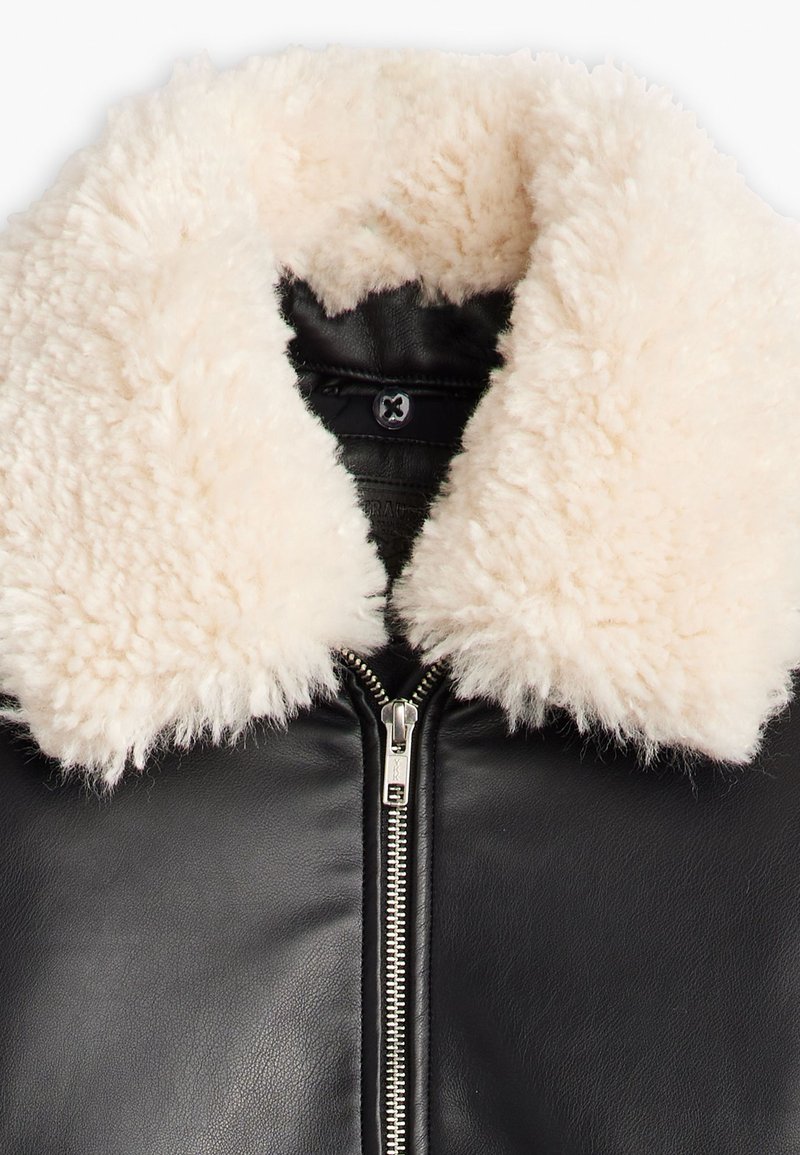 Chaqueta de cuero negra con un collar de piel sintética de color crema y detalles en cremallera plateada en la parte delantera, mostrando una textura suave y contraste.