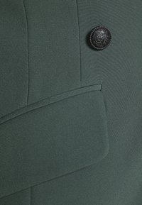 Temně zelená látka s hladkým povrchem; má černý dekorativní knoflík a detail kapsy s klopou na přední straně.