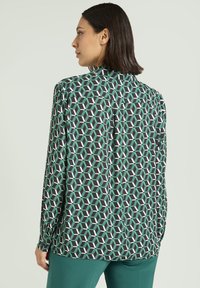 Koszula z długim rękawem w geometryczny wzór w kolorach czarnym, zielonym i białym, o luźnym kroju i pojedynczej plisie z tyłu, zestawiona z zielonymi spodniami.