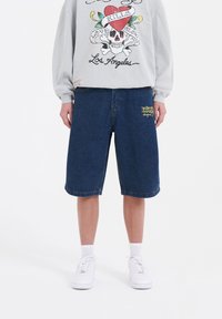 Tummansiniset denim-bermudashortsit rentona istuvana, keltaisella brodeeratulla logolla vasemmassa lahkeessa. Yhdistetty harmaaseen graafiseen huppariin.