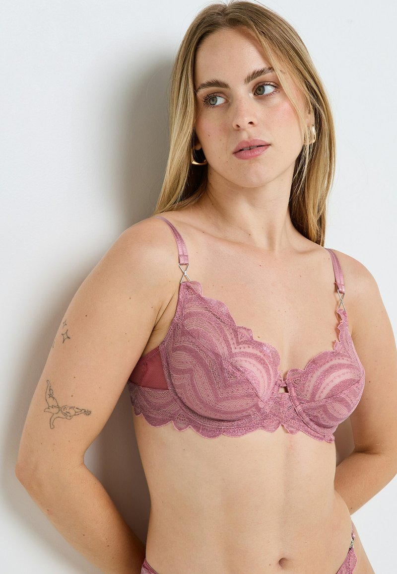 Soutien-gorge en dentelle de couleur rose pâle avec bords festonnés, motifs détaillés, armatures pour un bon maintien et bretelles réglables, orné d'un petit nœud central.