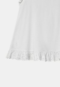 Biała bawełniana koszulka z krótkimi falbankowymi rękawami i wzorem haftu kwiatowego wzdłuż dolnego brzegu. Posiada logo w pobliżu brzegu.