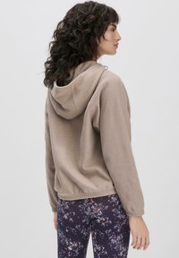 Felpa con cappuccio in un tenue color sabbia, dotata di tasca frontale, cappuccio regolabile e una texture morbida. Abbinata a leggings scuri con motivi floreali.
