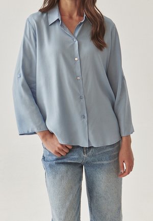 Kobieta ubrana w jasnoniebieską, rozpinaną bluzkę z długim rękawem, luźno wpuszczoną w wyblakłe niebieskie dżinsy, stojąca z jedną ręką w kieszeni.