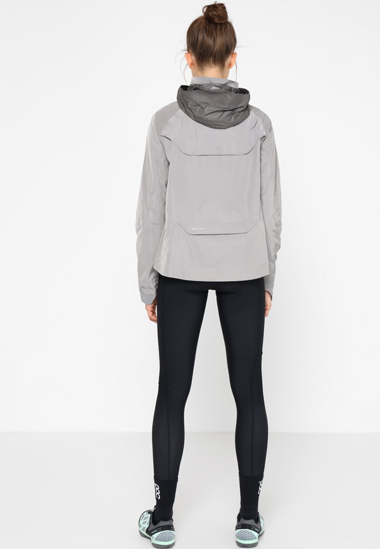 Hellgraue Jacke mit Kapuze, die eine glatte Textur und eine Gesäßtasche aufweist. Kombiniert mit schwarzen Leggings und Schuhwerk mit tealfarbenen Akzenten.