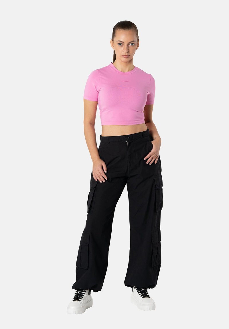 Nainen seisoo käsi mustien cargo-housujen taskussa, pukeutuneena vaaleanpunaiseen lyhytpaitaiseen t-paitaan ja valkoisiin korokepohjakenkiin valkoista taustaa vasten.