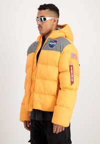 Chaqueta de plumas naranja brillante con hombros grises, logo de NASA, parche de la bandera estadounidense y detalles en rojo. Cierre frontal con cremallera, corte holgado.