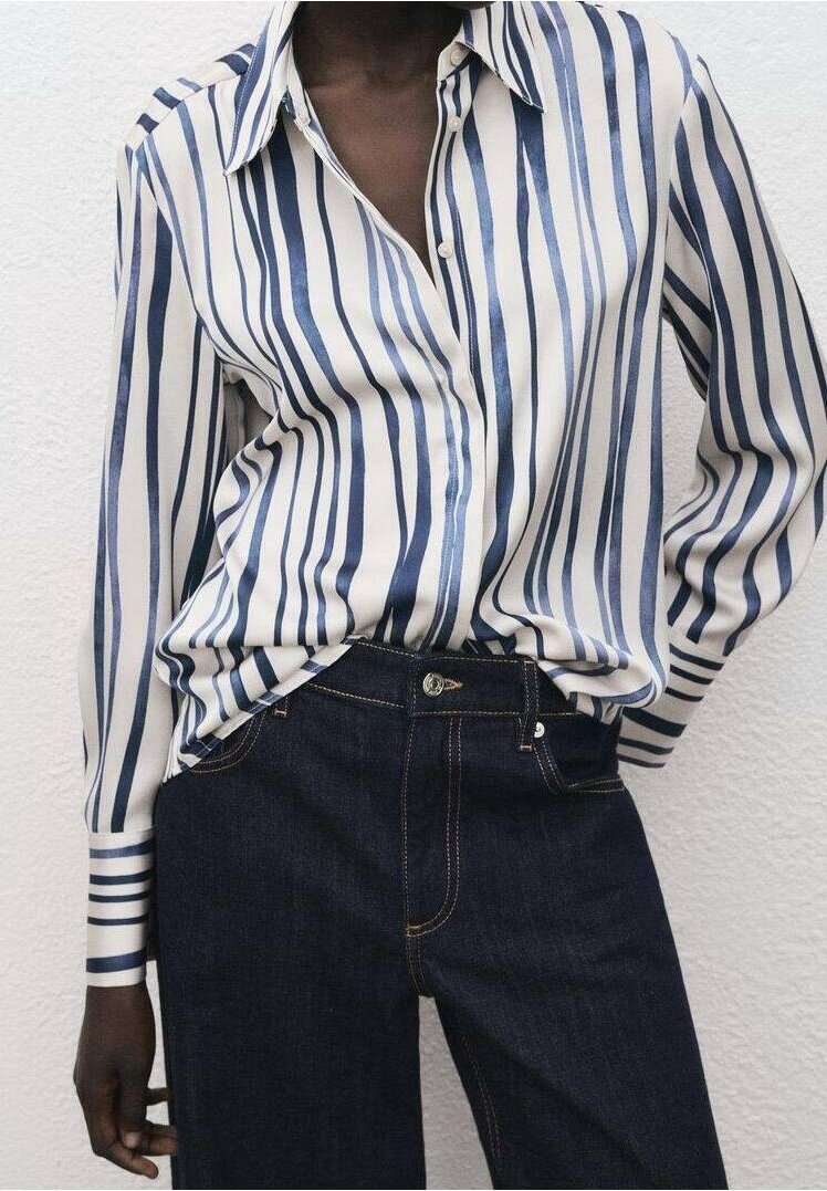 Personne portant une chemise blanche à rayures verticales bleu marine rentrée dans un jean taille haute bleu foncé contre un mur blanc texturé.