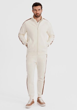 Man staat met handen in zijn zakken, gekleed in een crèmekleurig trainingspak met bruine zijstrepen en beige sneakers, tegen een witte achtergrond.