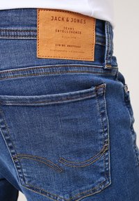 Los jeans de mezclilla azul presentan un parche de cuero visible etiquetado "JACK & JONES" en la parte trasera, con costuras detalladas y diseño de bolsillo.