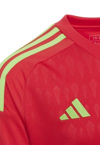 Maillot de sport rouge avec trois rayures vert clair sur les épaules et un logo Adidas brodé en vert foncé, tissu avec un motif en diamant texturé.