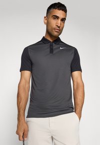 Nike polo krekls tumši pelēkā krāsā ar īsām piedurknēm, klasisku apkakli un trīs pogu slēgšanu; uz krūtīm ir neliels balts logotips.