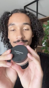 Conteneur de poudre noir en forme circulaire tenu dans les mains, avec un fond transparent, une texture lisse et un logo métallique sur le dessus.