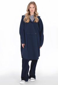Tengerészkék vízálló esőkabát kapucnival, laza illeszkedéssel, elöl gombos záródással, egyetlen mellzsebbel, csíkos ing és sötét farmer fölött viselve.