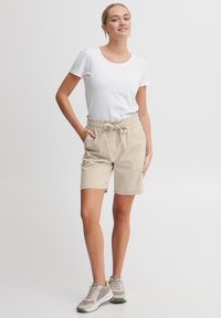 Shorts beiges à taille élastique avec poches latérales, associés à un t-shirt blanc à col rond et des baskets grises. Tissu lisse avec une coupe décontractée.