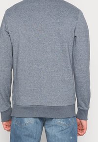 Hombre vistiendo un suéter liso de manga larga gris medio y unos jeans de denim azul claro, visto desde atrás.