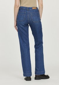 Jeans en denim bleu foncé à coupe droite, avec deux poches arrière et une étiquette en cuir sur la ceinture. Détail de couture au niveau des ourlets.