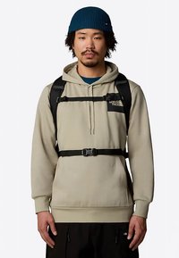 Beige hoodie met een voorzak, zwarte verstelbare bandjes en een logo. Draagt over een teal kleurig shirt en gecombineerd met zwarte broek, voorzien van een ingewikkelde gebreide beanie.