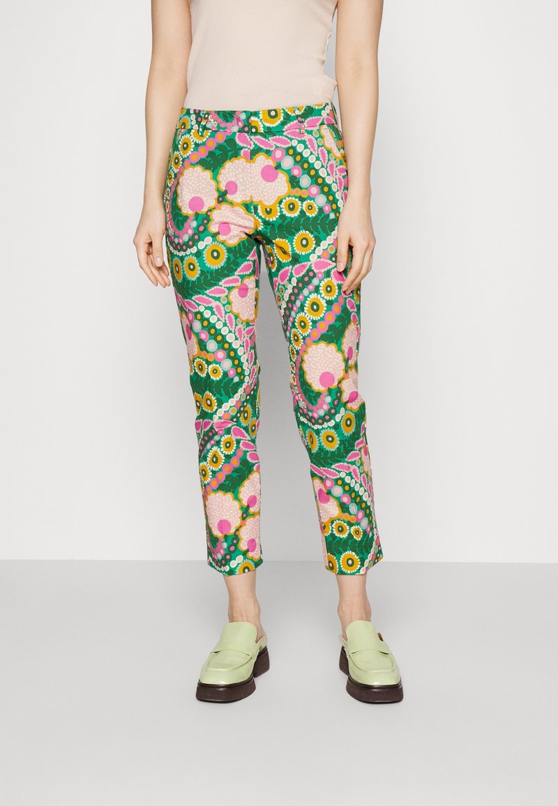 Femme portant un pantalon à motifs floraux verts, roses et jaunes avec des chaussures slip-on vert clair, debout sur un fond blanc uni.