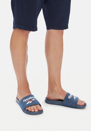 Osoba ubrana w granatowe szorty i niebieskie klapki Reebok, stojąca na białym tle.