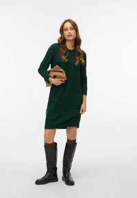 Zelené pletené šaty s dlouhými rukávy, volný střih, nošeny s černými kozačkami po kolena a držící tanovou kabelku. Jednoduchá textura, žádné vzory.