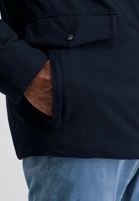 Person mit einer dunklen Jacke, die teilweise die Hand in der Seitentasche hat, über hellblauen Cordhosen vor einem schlichten Hintergrund.