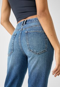 Detailní záběr na osobu, která má na sobě modré džíny s vysokým pasem a černý crop top, přičemž její pravá ruka je uvolněně položená podél těla.