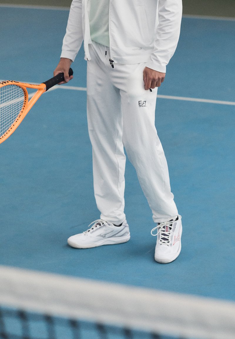Survêtement de sport blanc en tissu texturé, tenant une raquette de tennis orange, associé à des baskets blanches avec des accents noirs.