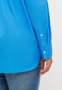 Langermet blå skjorte med en jevn tekstur, med knapper ved mansjettene og en lett buet hem. Brukt med denimjeans.