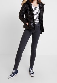 Chaqueta de plumas negra con capucha forrada de piel, camiseta de rayas blanca y negra de manga larga, jeans ajustados gris oscuro y zapatillas altas azules.