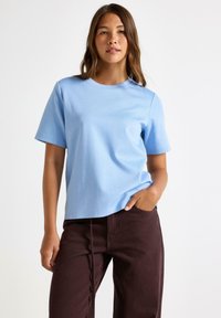 Jongeman met lang bruin haar draagt een lichtblauw T-shirt met korte mouwen en donkerbruine broek, staande tegen een simpele witte achtergrond.